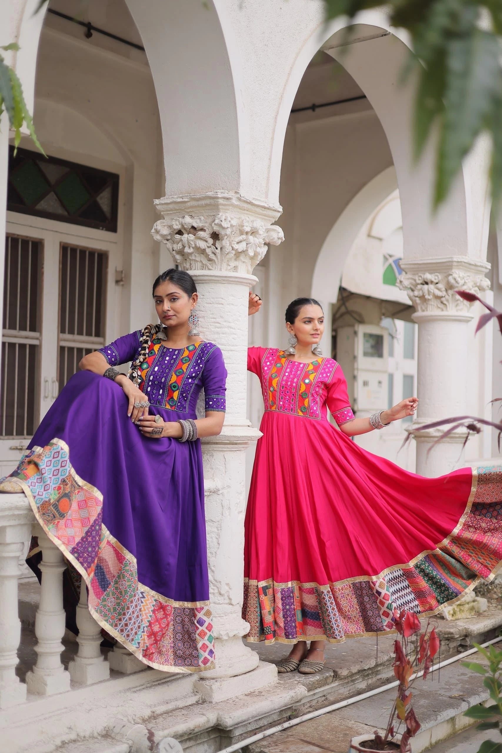 Two women in colorful traditional dresses standing in front of a white architectural background.